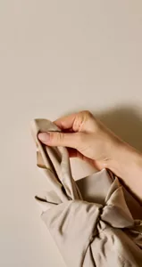 Close up of person's hand tying knot of beige Furoshiki wrapping on top of a beige surface